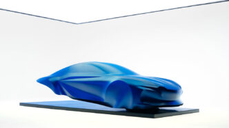 Yeni Alpine A110'un tasarımını önizleyen mavi örtülü heykel, ön 3/4 görünüm